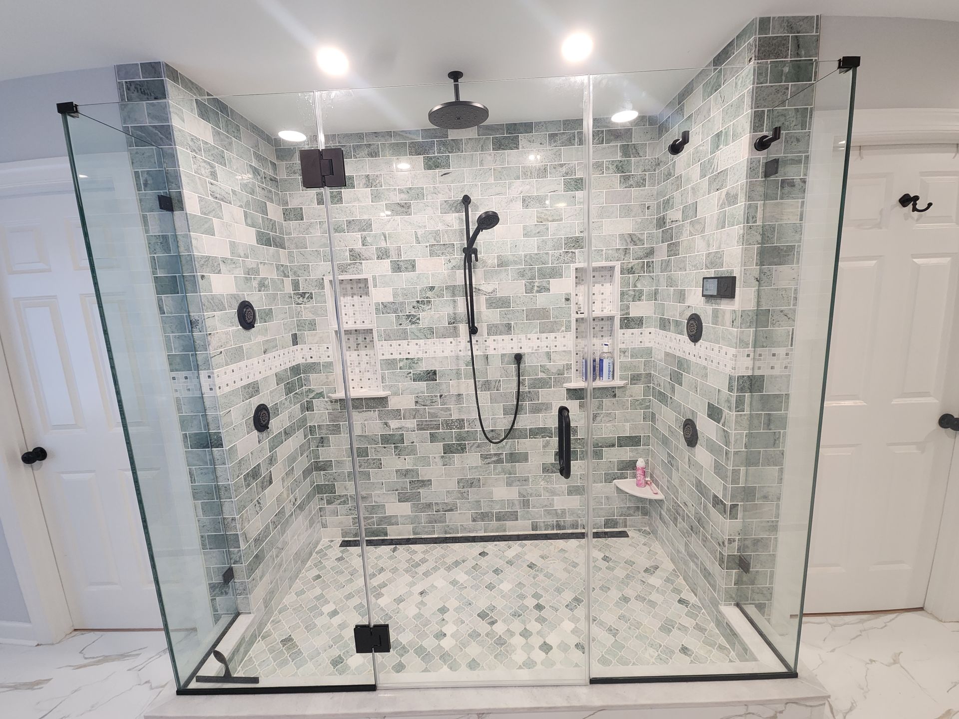 Walk-in shower with green tiled walls, glass doors, black fixtures, and mosaic floor.