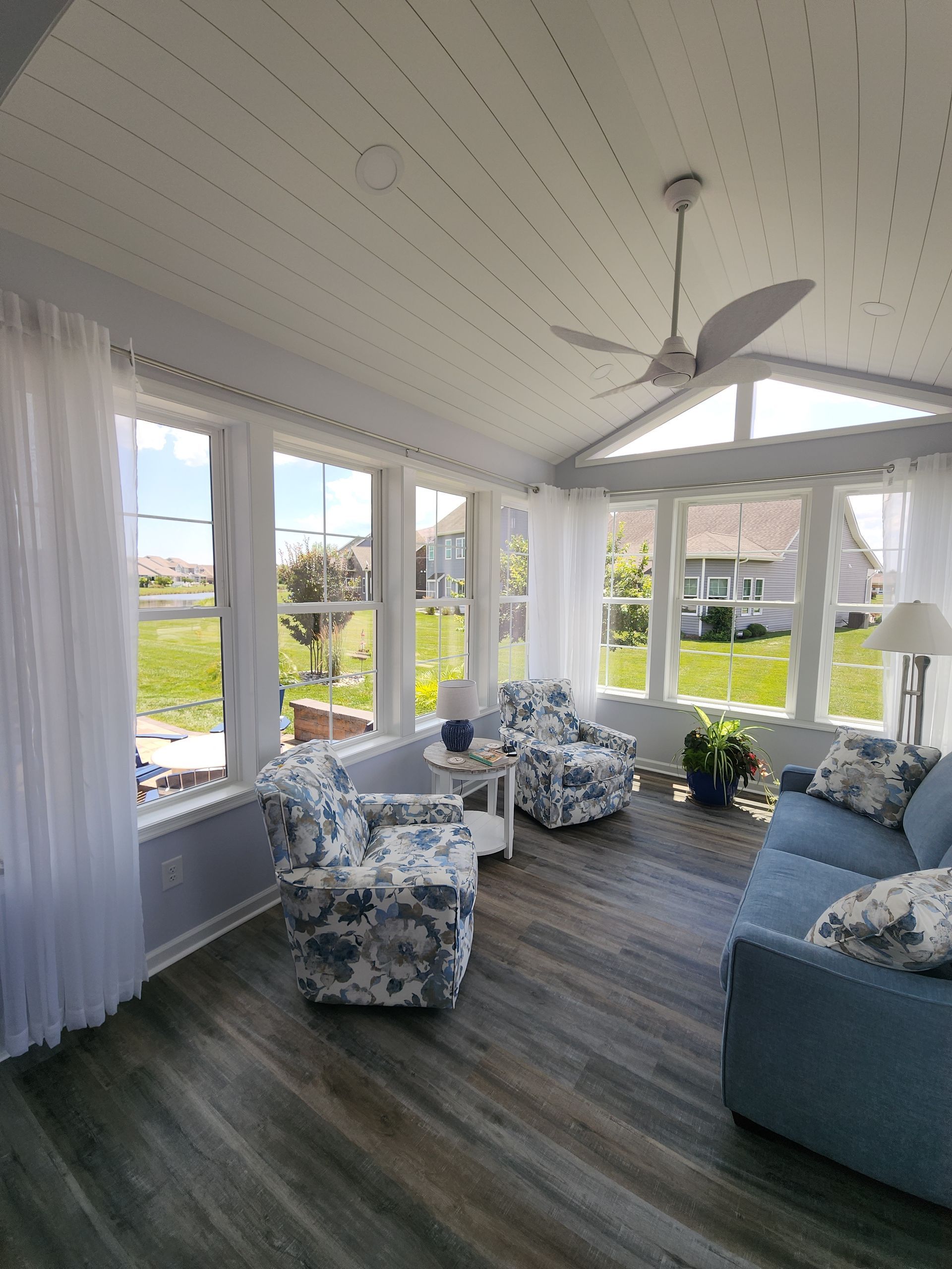Sunroom with blue and white floral armchairs, sofa, and sheer curtains. Wooden floors, white walls, and ceiling fan.