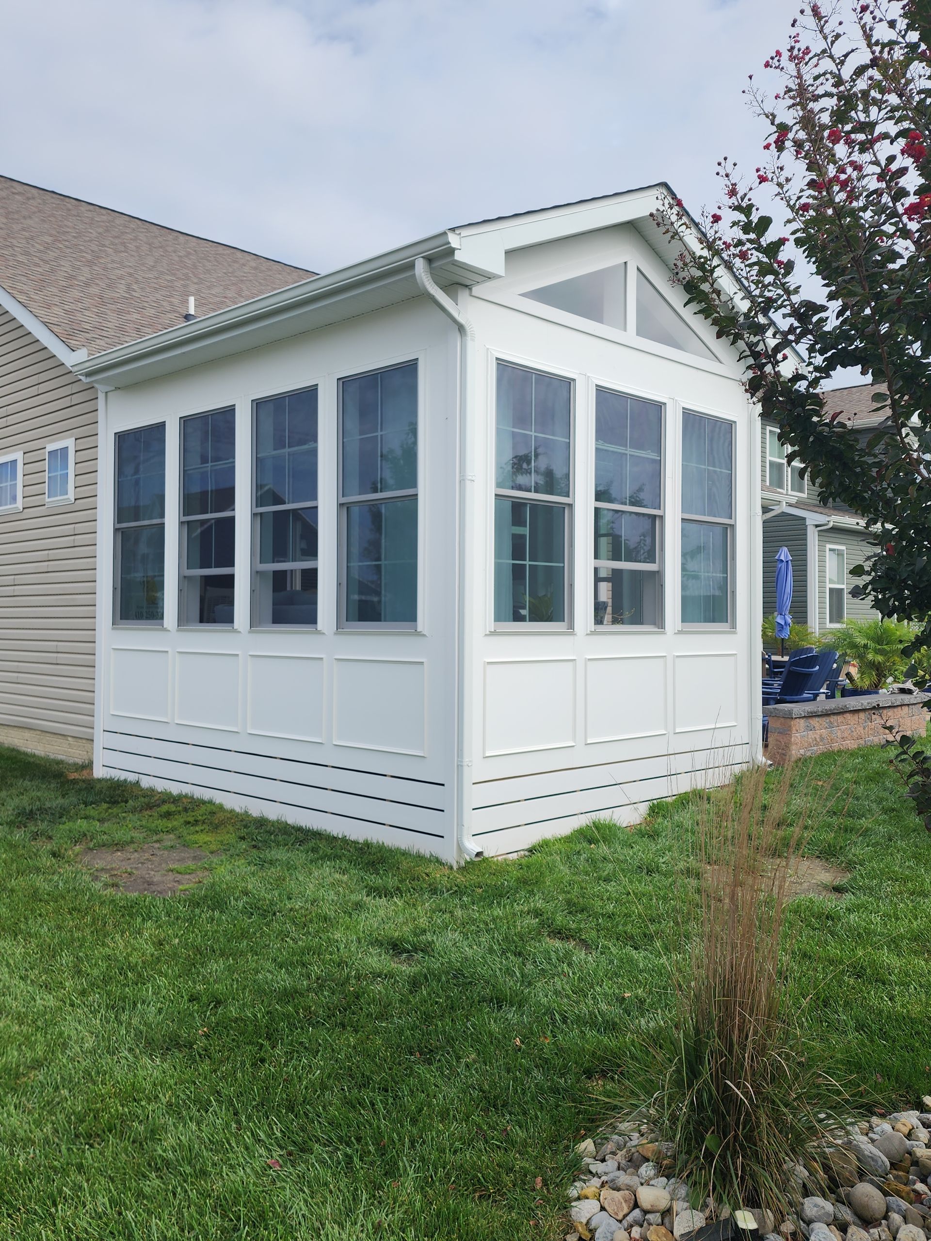 White sunroom addition with windows and gabled roof, attached to a light-colored brick house, green yard.