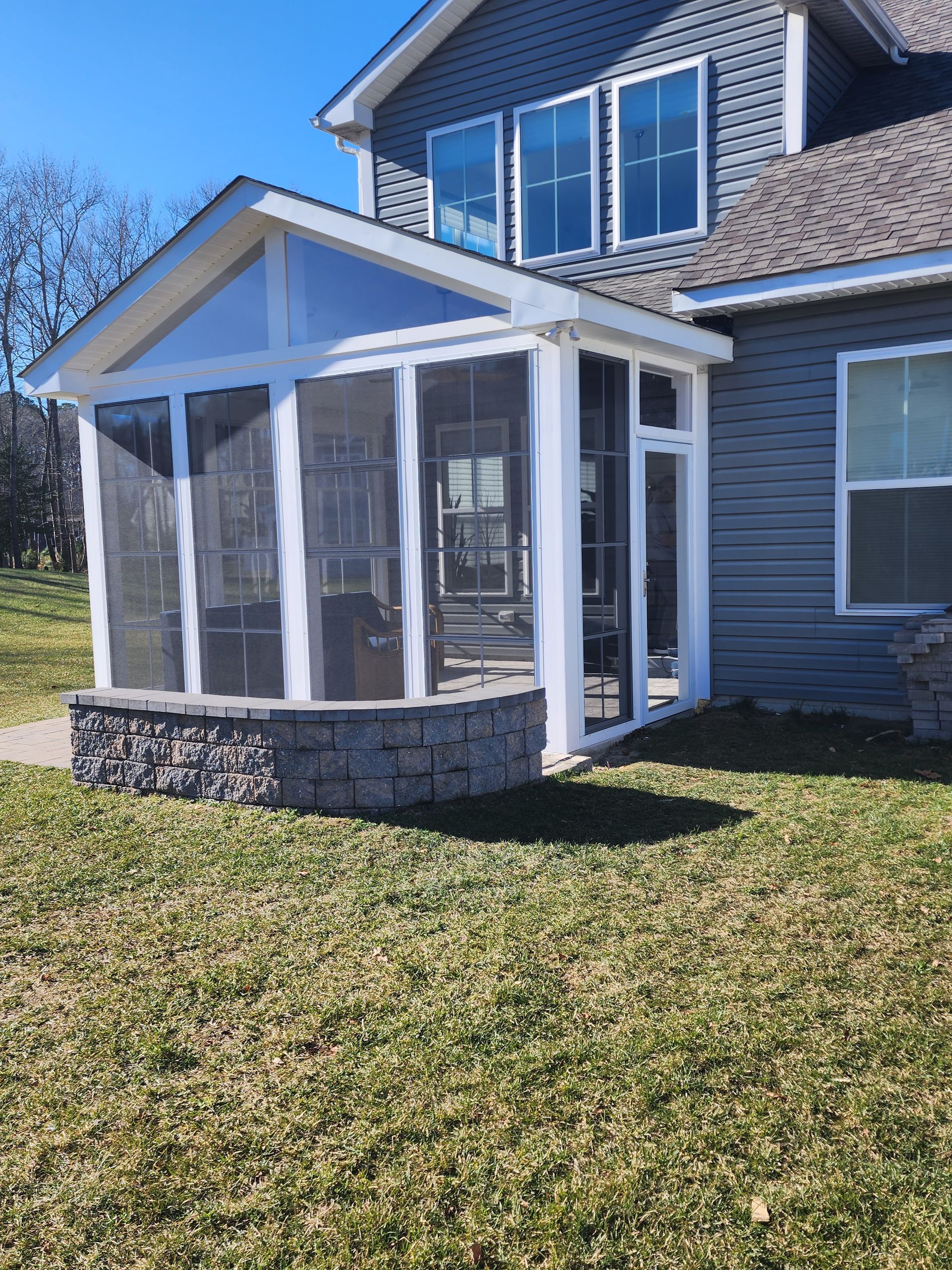 Screened-in porch attached to a blue house with stone base. White trim, windows, and green lawn.