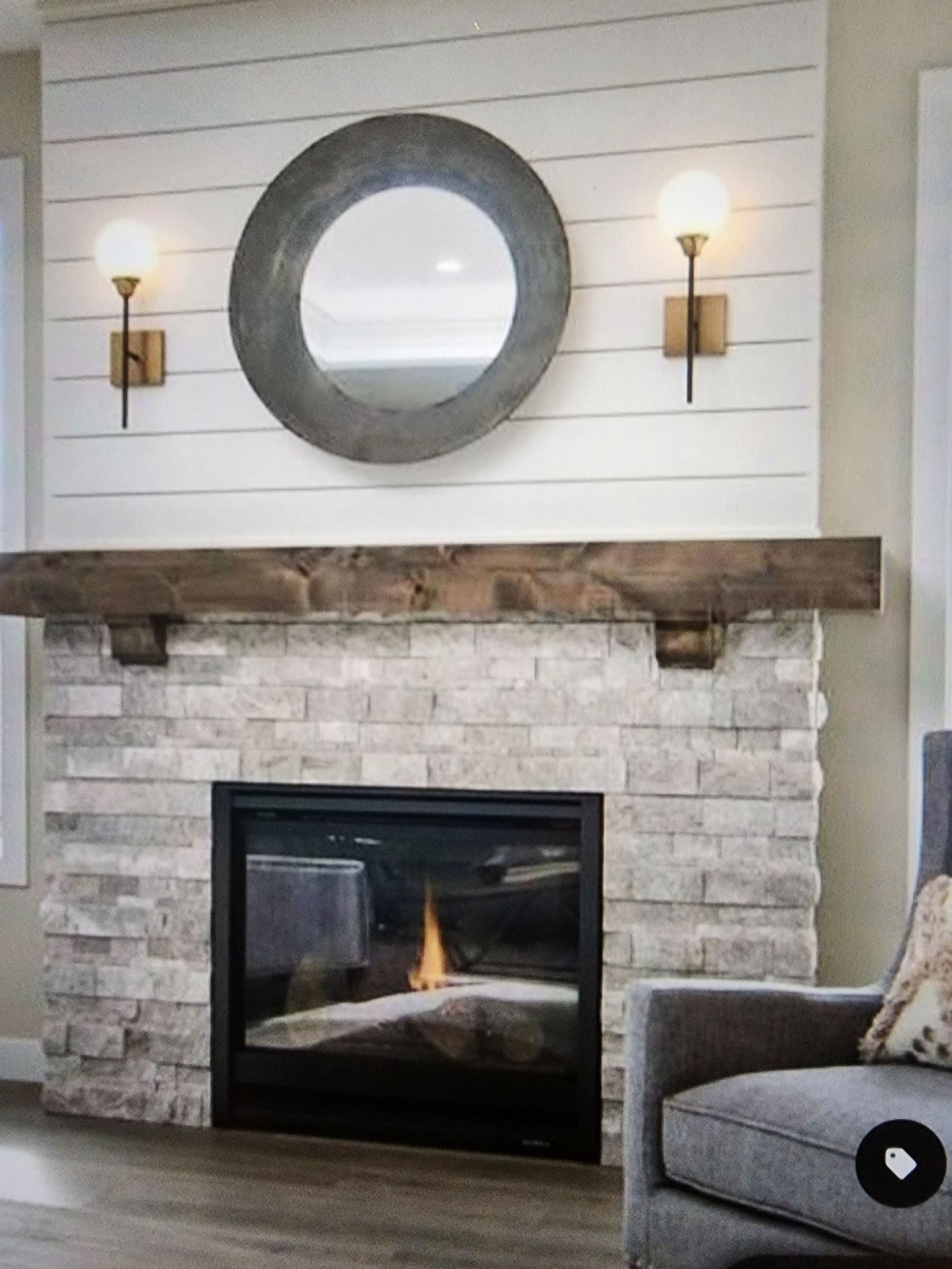 Fireplace with stone facade, dark wood mantel, round mirror, sconces, and white shiplap wall.