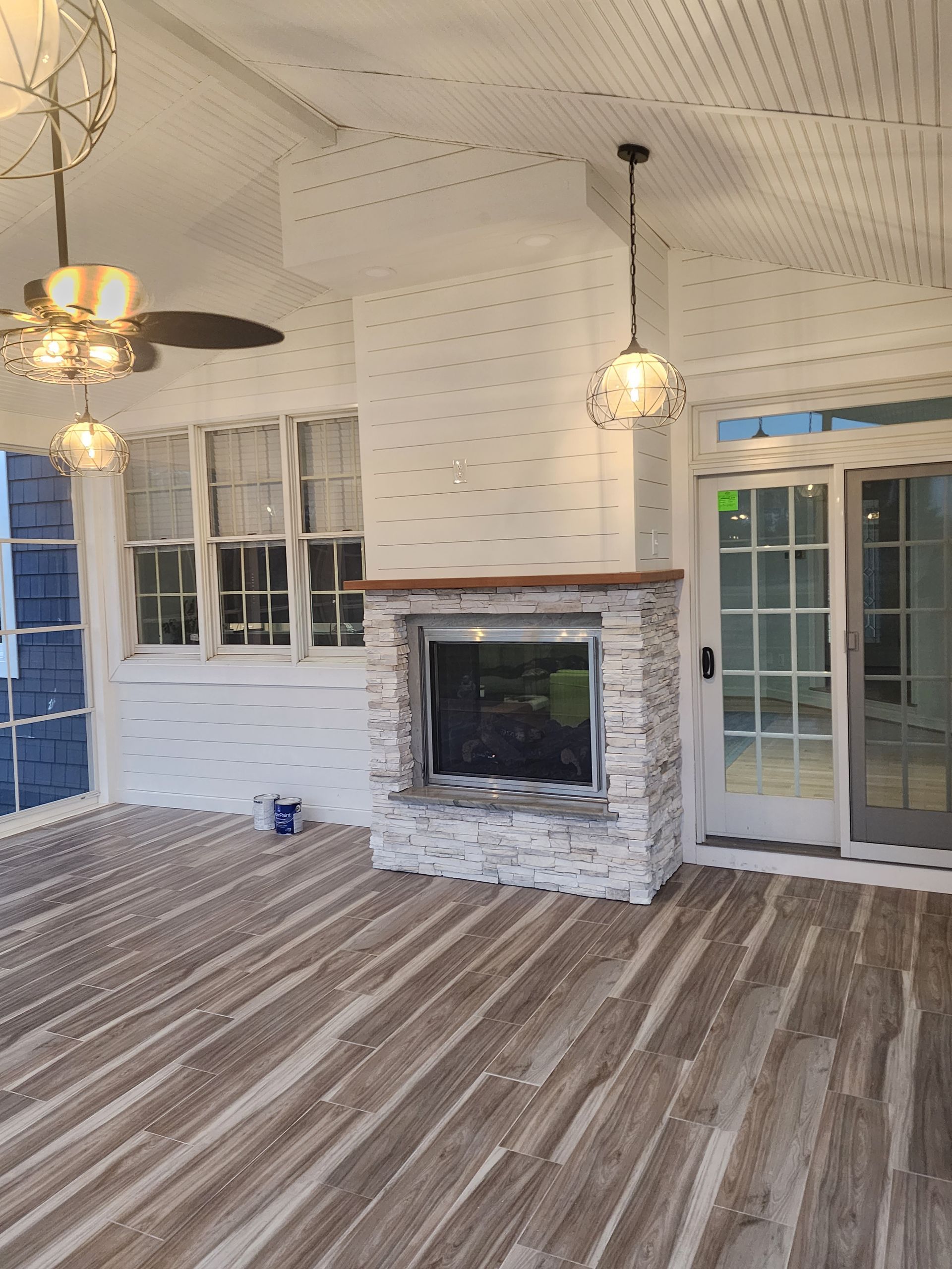 A sunroom with a fireplace, ceiling fan, and sliding doors.  White brick and light wood flooring.