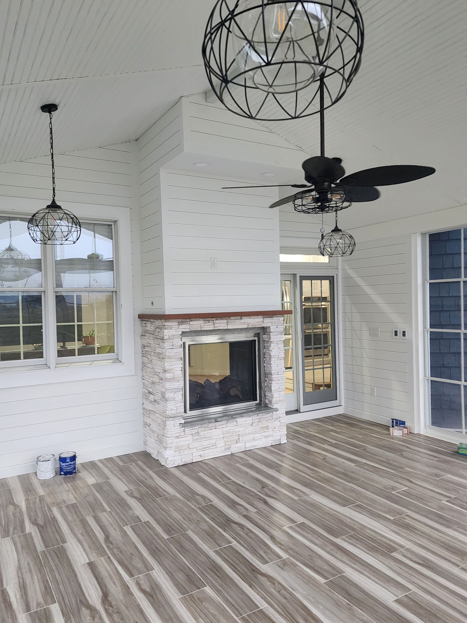 Covered outdoor room with fireplace, wood-look flooring, white walls, and decorative light fixtures.
