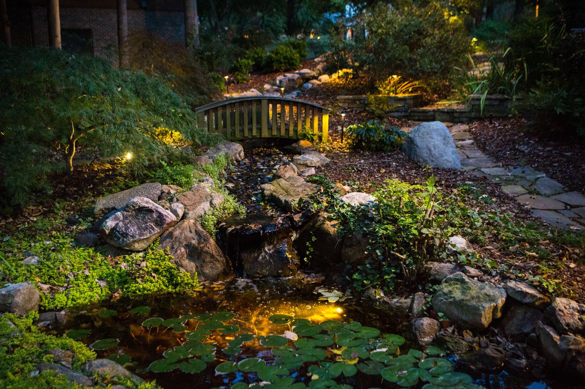 Lit garden scene at dusk; small wooden bridge over a pond, rocky terrain with green foliage and lights.