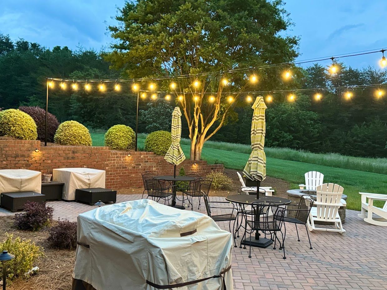 Patio with string lights, tables, chairs, and covered furniture on a brick surface at dusk.
