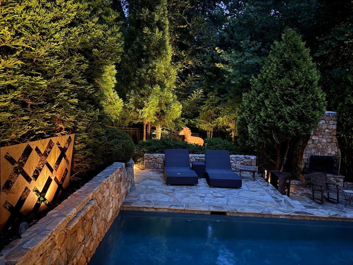 Poolside lounge area at night, illuminated by soft lighting. Stone patio, pool, trees.