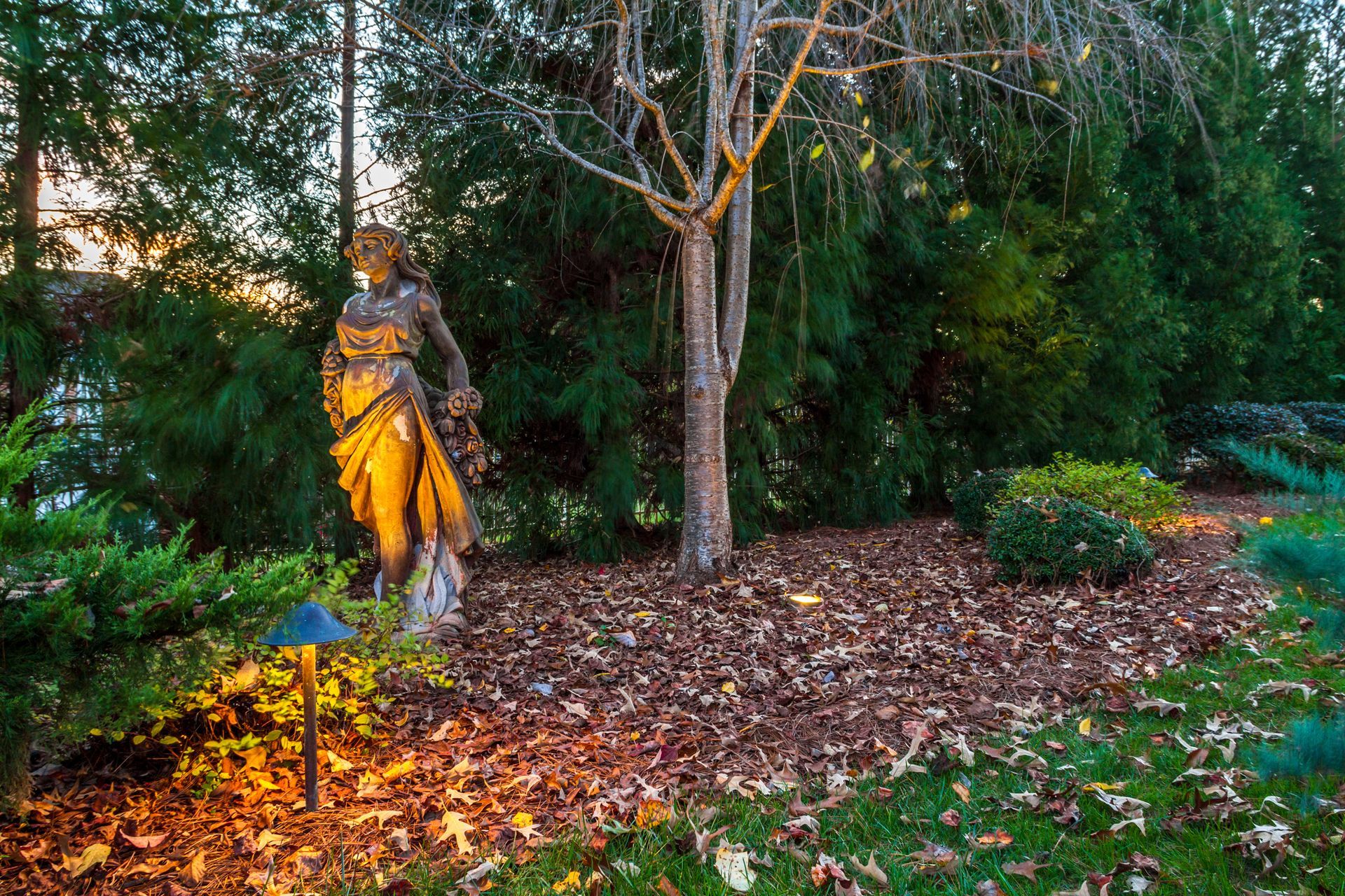 Bronze statue in a garden, lit by a spotlight, surrounded by foliage and fallen leaves.