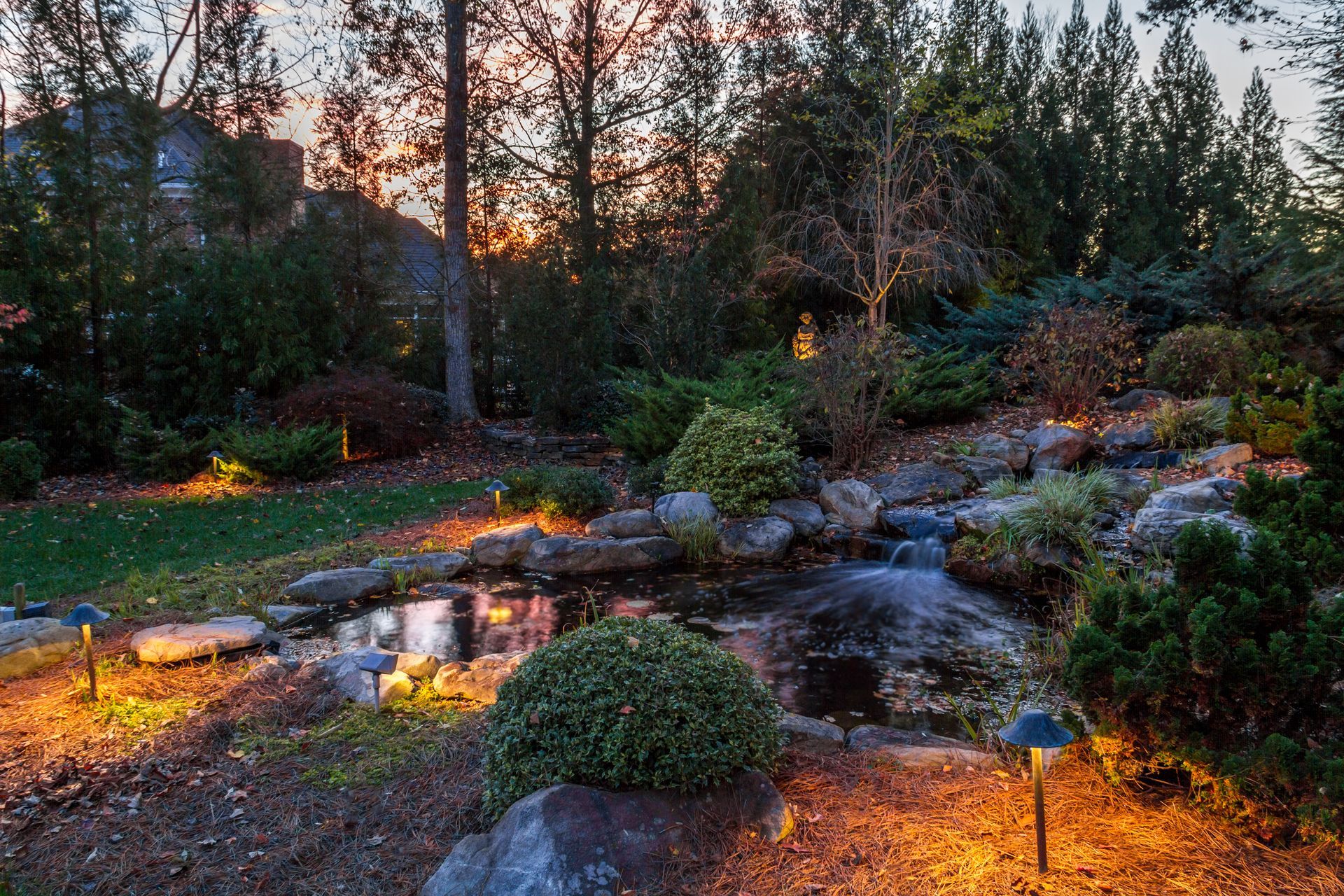 Landscape with pond and waterfall at dusk, illuminated by warm lights; trees in the background.