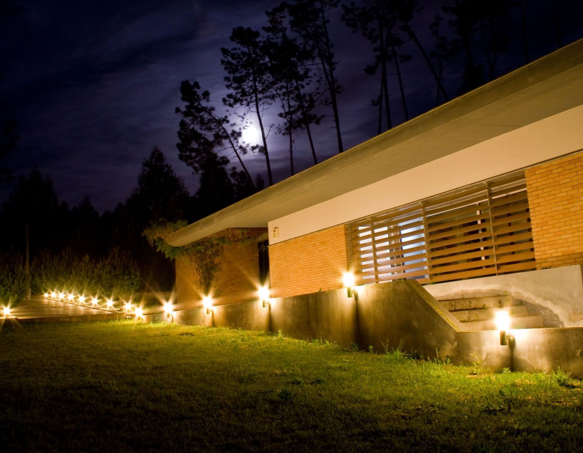 Modern house at night, lit pathway and building lights against a dark sky with a moon.