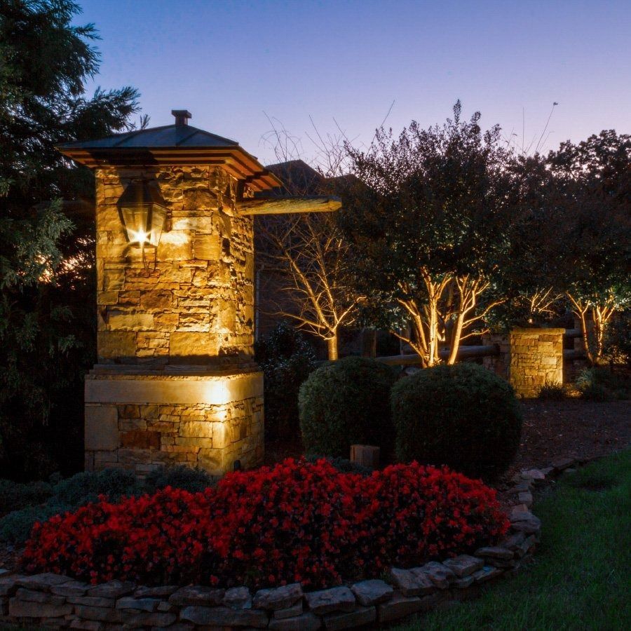 Stone entry pillars with illuminated landscape, shrubs, and trees at dusk.