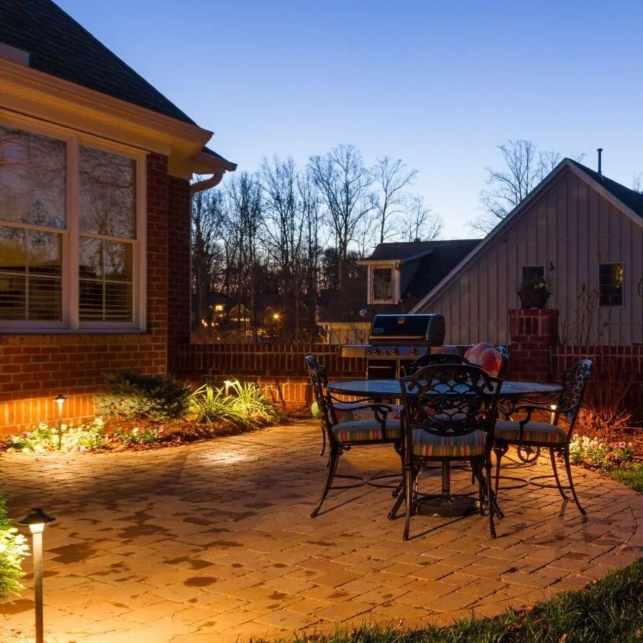 Brick patio with outdoor table and chairs illuminated by warm lighting at dusk.