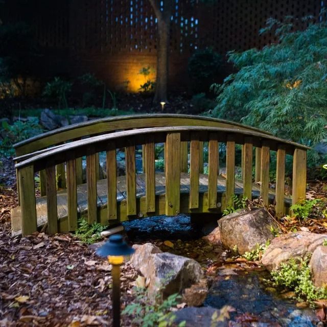 Wooden footbridge over a stream in a garden, lit at night.