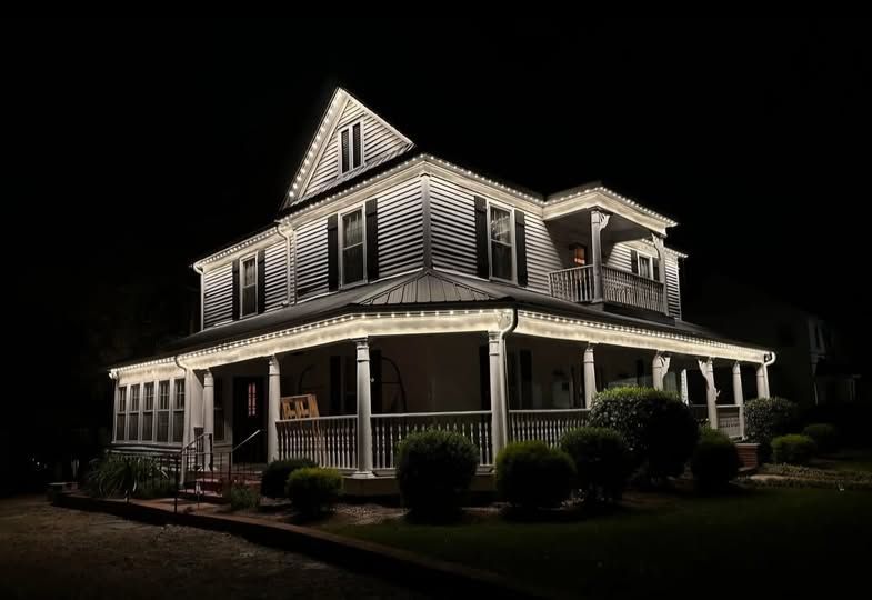 Lit-up Victorian house at night, white trim, porch, bushes, and a dark sky.
