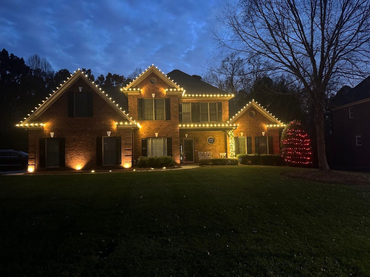 Two-story brick house illuminated with warm white Christmas lights along the roofline, with a red Christmas tree on the side.