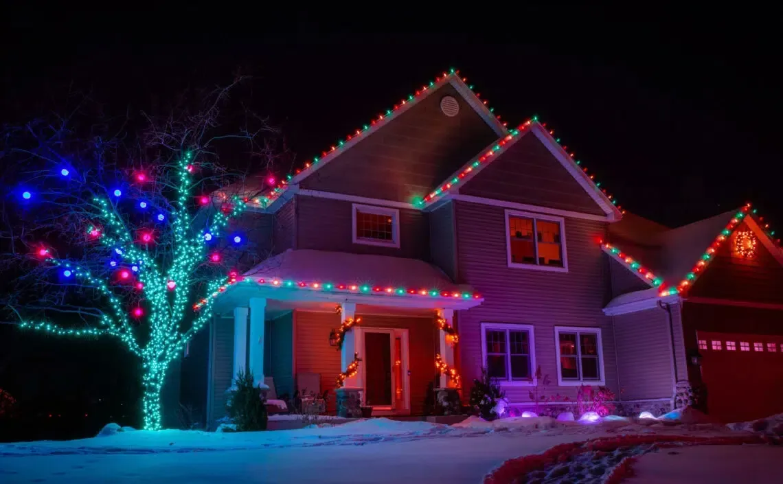 House decorated with colorful Christmas lights against a dark night sky, snow on the ground.