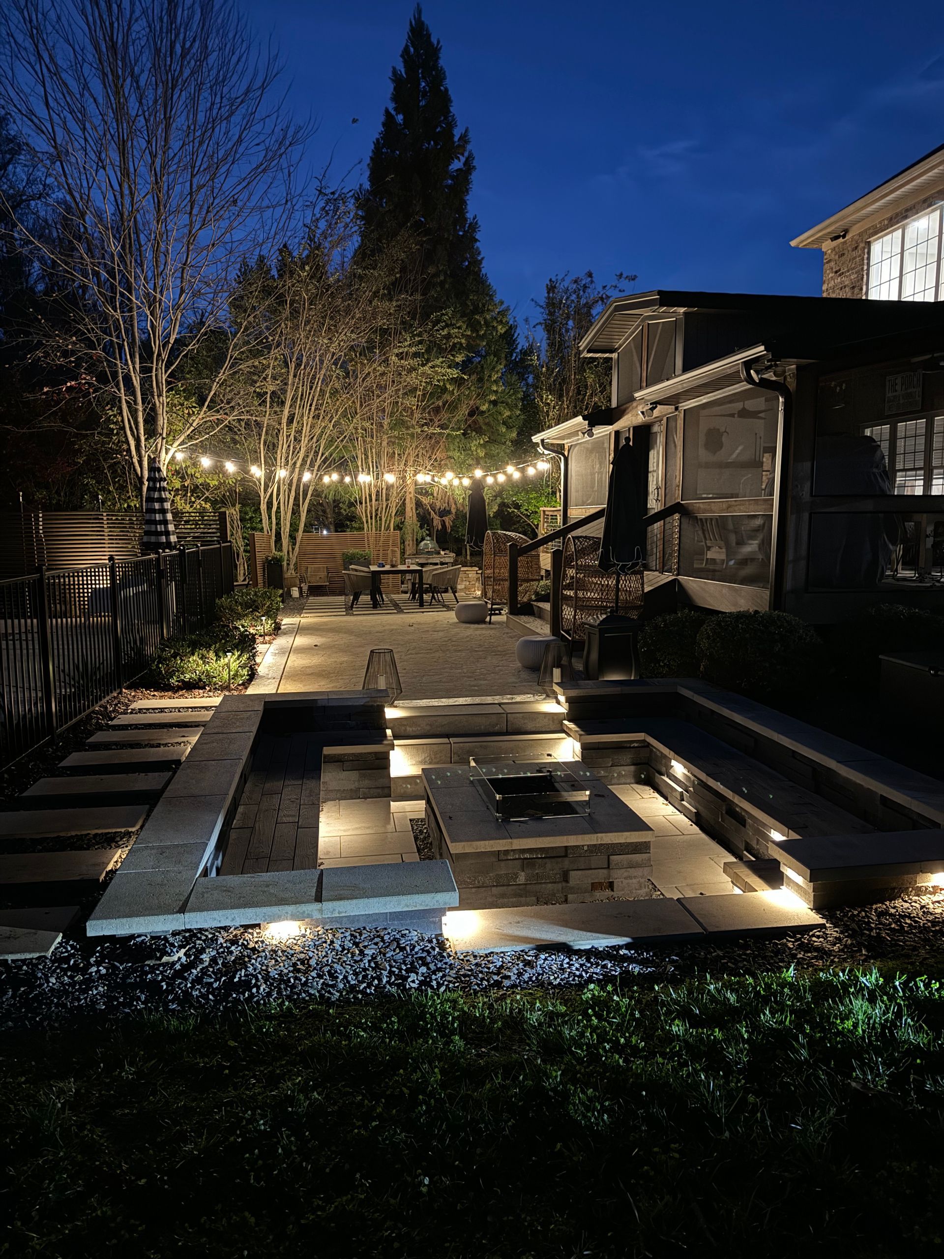 Nighttime backyard patio with string lights, fire pit, stone steps, and house.