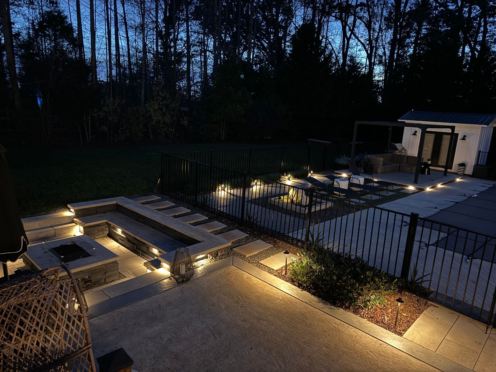 Outdoor patio at dusk, with fire pit, seating, and lit pathways. Trees in background, dark sky.