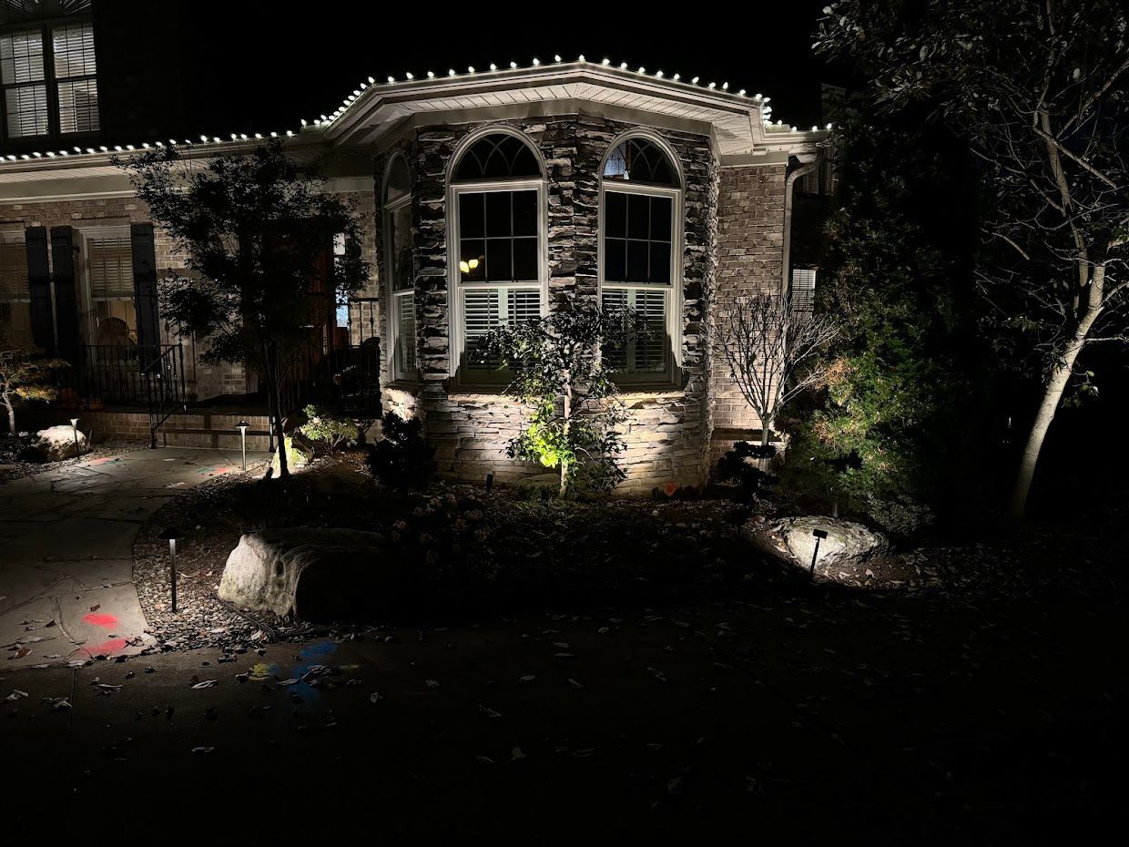 Night view of a house with stone facade, arched windows, and landscape lighting illuminating trees and bushes.