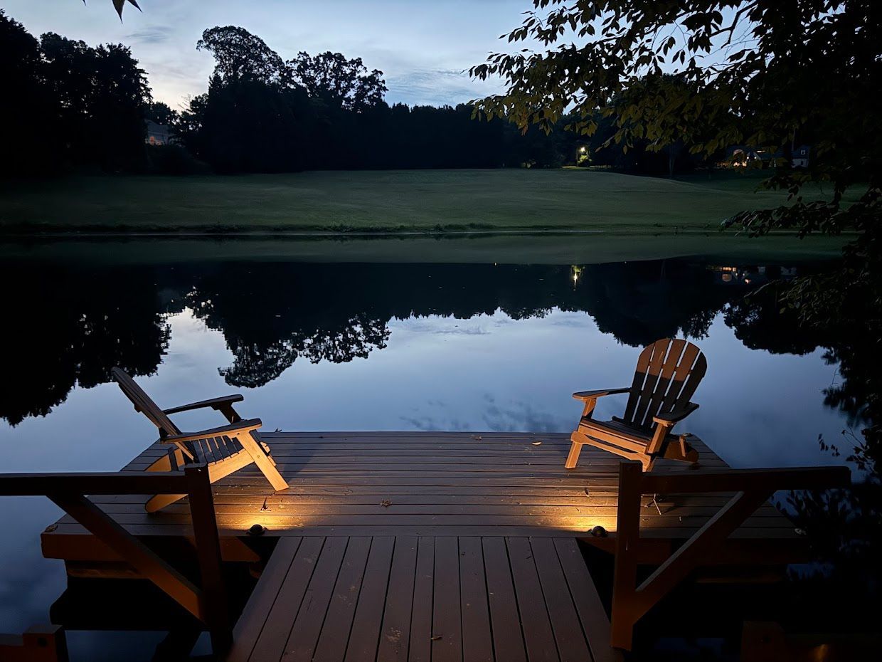 Two wooden chairs on a dock over a calm lake at dusk, with ambient lighting and trees in the background.