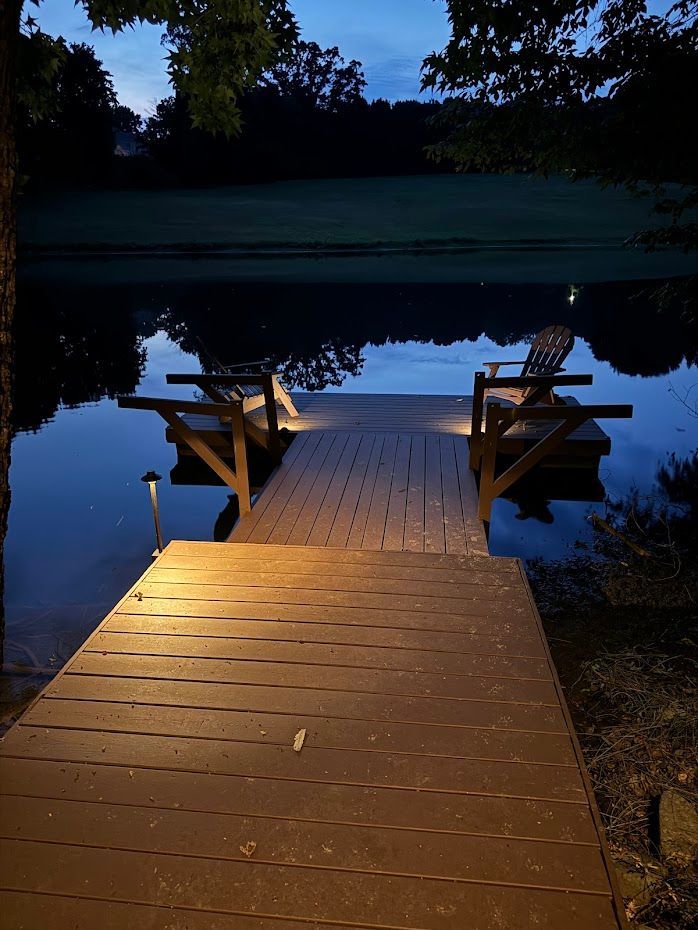 Wooden dock with two chairs illuminated by lights, extending into calm water at dusk.