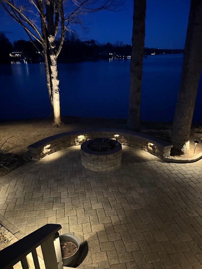 Nighttime outdoor patio with a fire pit, overlooking a dark lake, lit by warm lights.