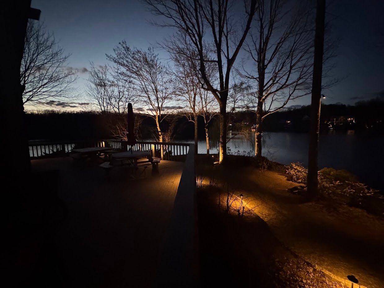 Evening scene by a lake. Warm spotlights illuminate a dock and trees under a twilight sky.