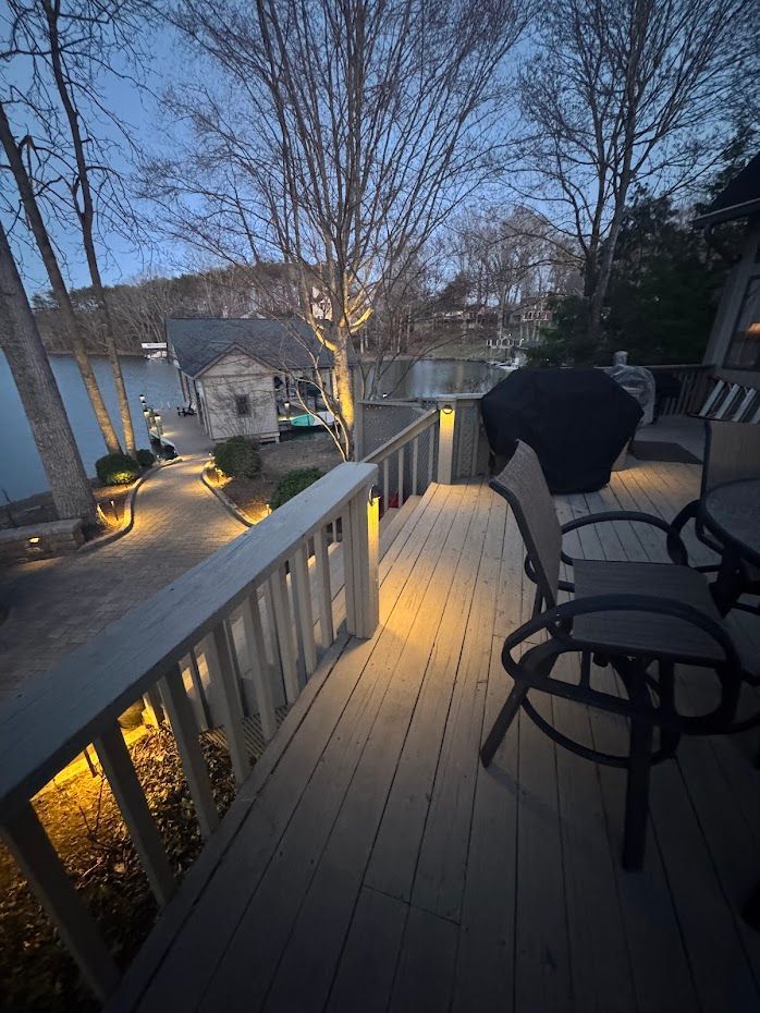 A deck lit by warm lights overlooks a lake at dusk. A grill and chairs are in the foreground.