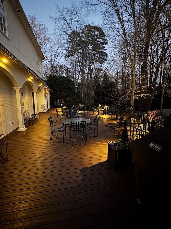 A wooden deck with patio furniture lit by warm lights at dusk, beside a white building and trees.