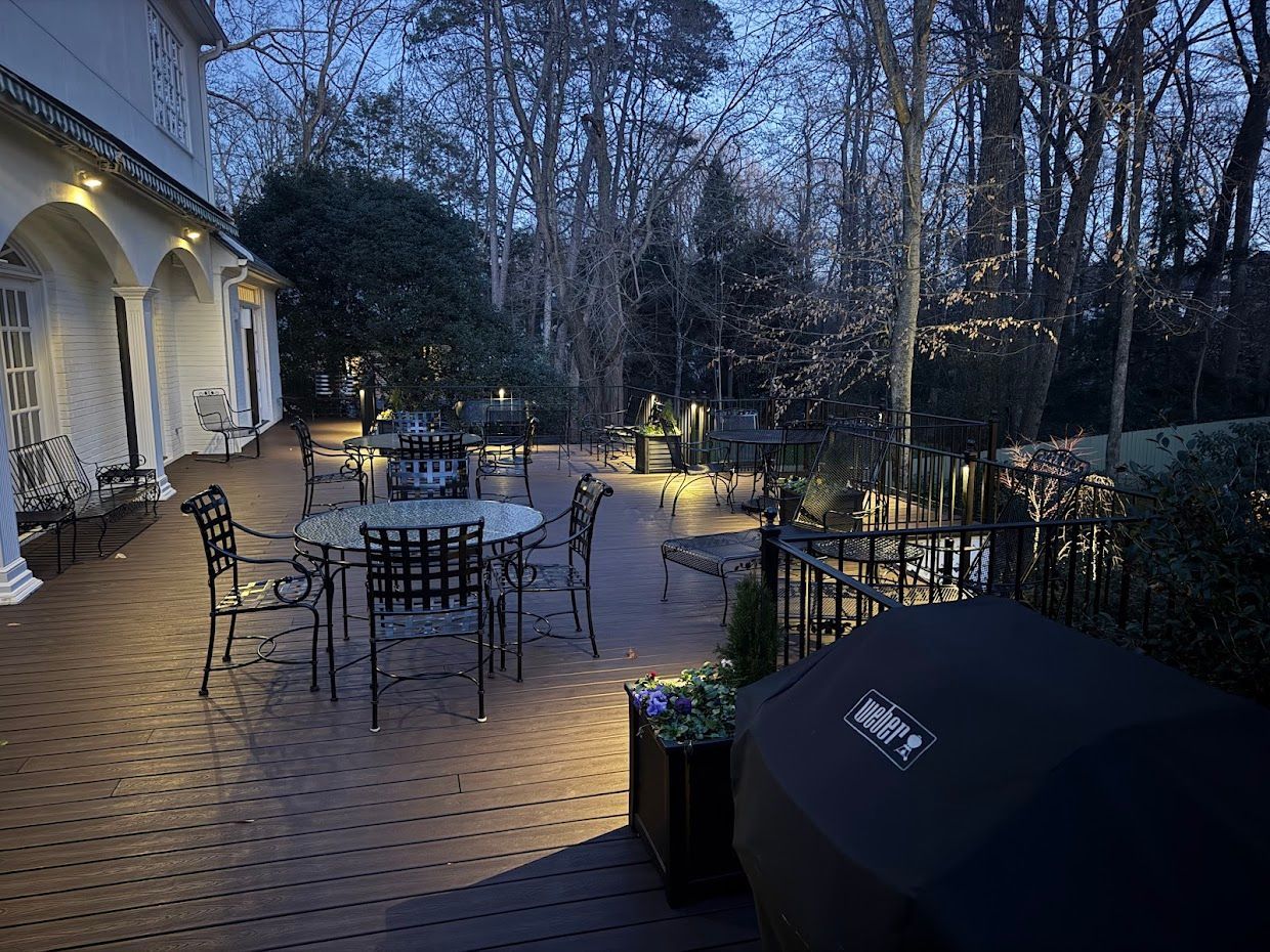 Outdoor patio at dusk, with round tables, chairs, and a grill. Lights illuminate the deck and surrounding trees.
