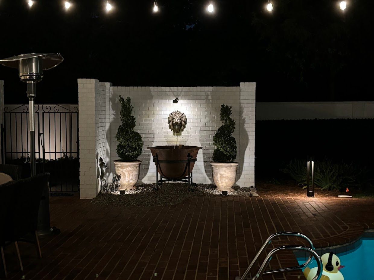 Nighttime backyard scene with lit patio, wall with planters, and pool. String lights overhead.