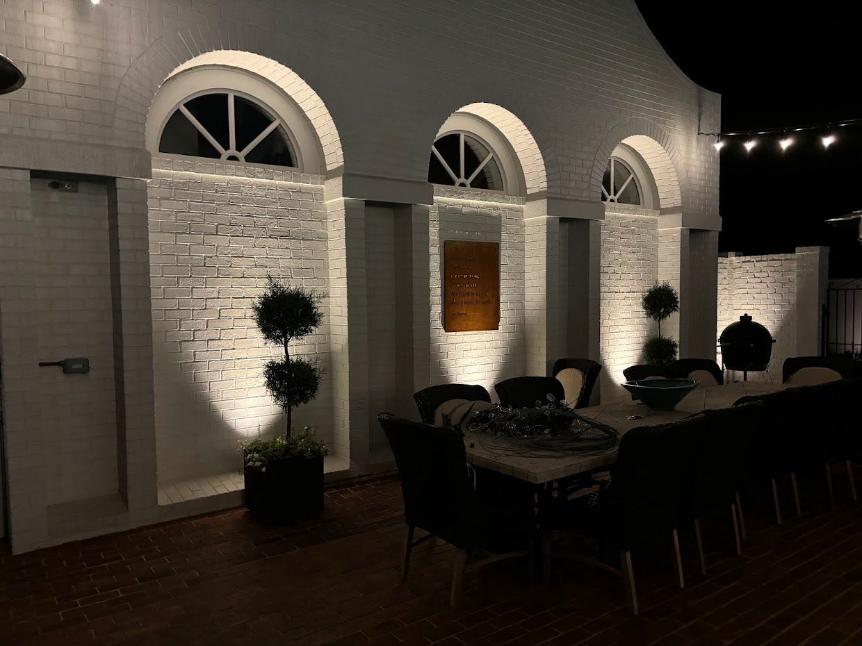 Outdoor dining area with white arches, brick wall, and a long table set for a meal.