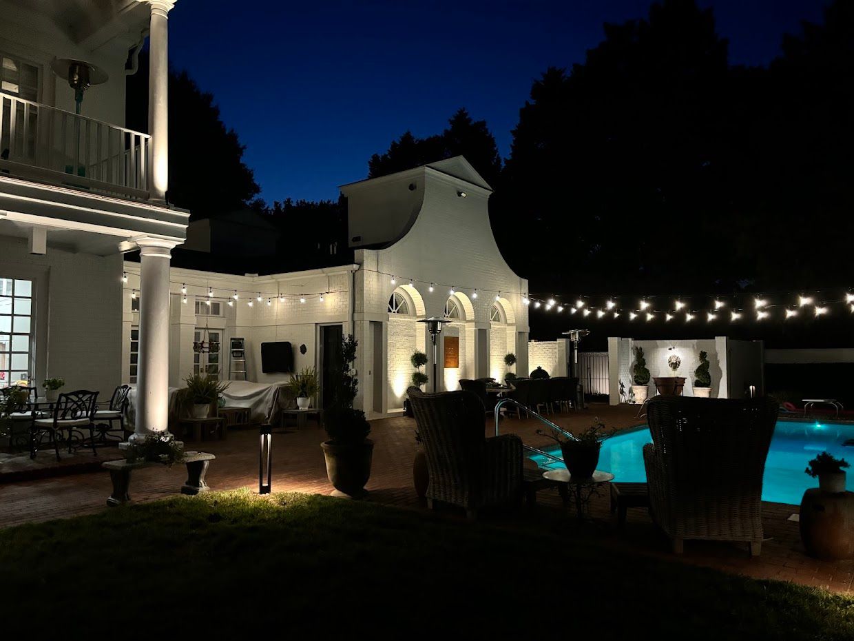 Night view of a lit backyard with a pool, patio furniture, and a white building. String lights and uplighting.