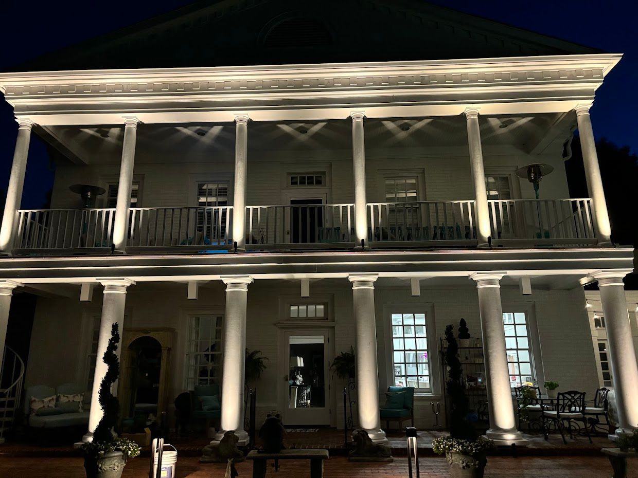 Lit, two-story white house with columns, balcony, and green roof at night.