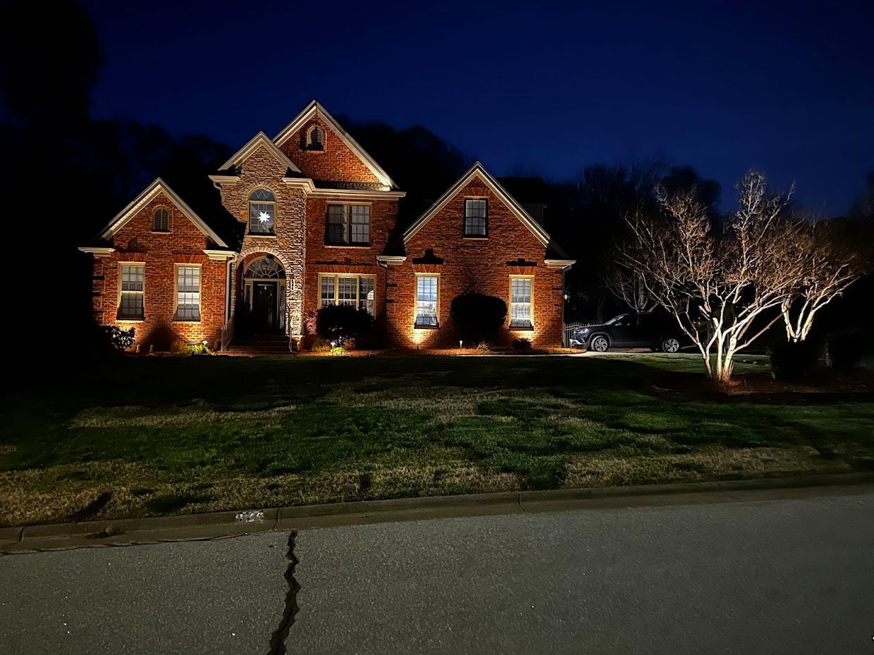 Brick house illuminated at night with spotlights, tree lit up to the right, front yard, dark sky.