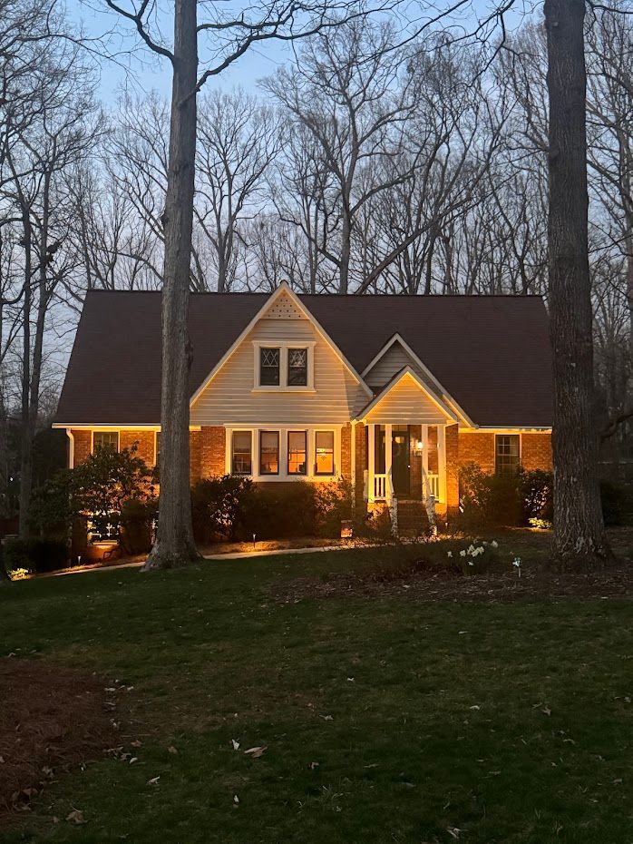 A house at dusk with warm exterior lights, surrounded by trees and a green lawn.