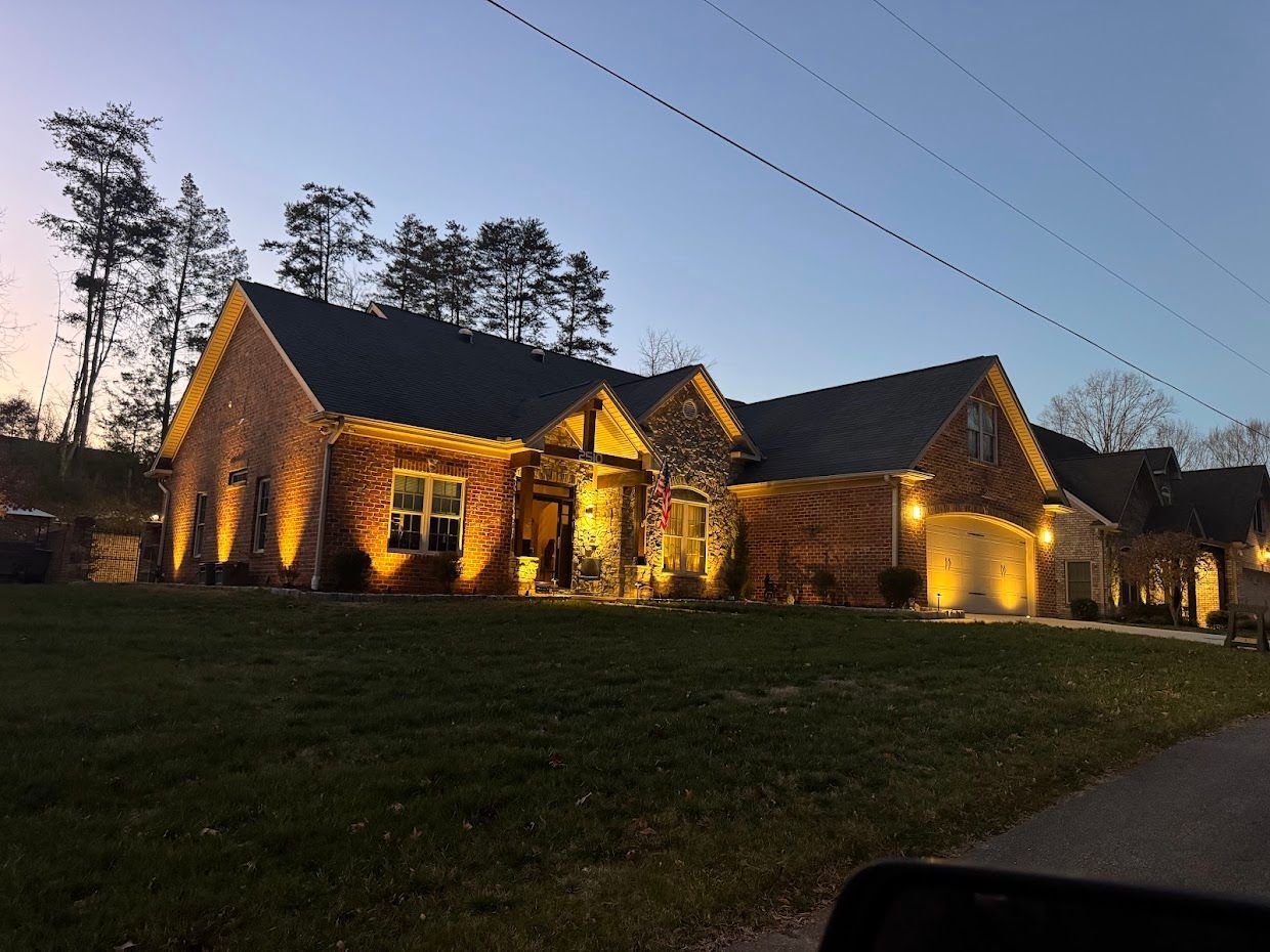 Brick house at dusk, lit with warm yellow lights. Trees and power lines are visible in the background.