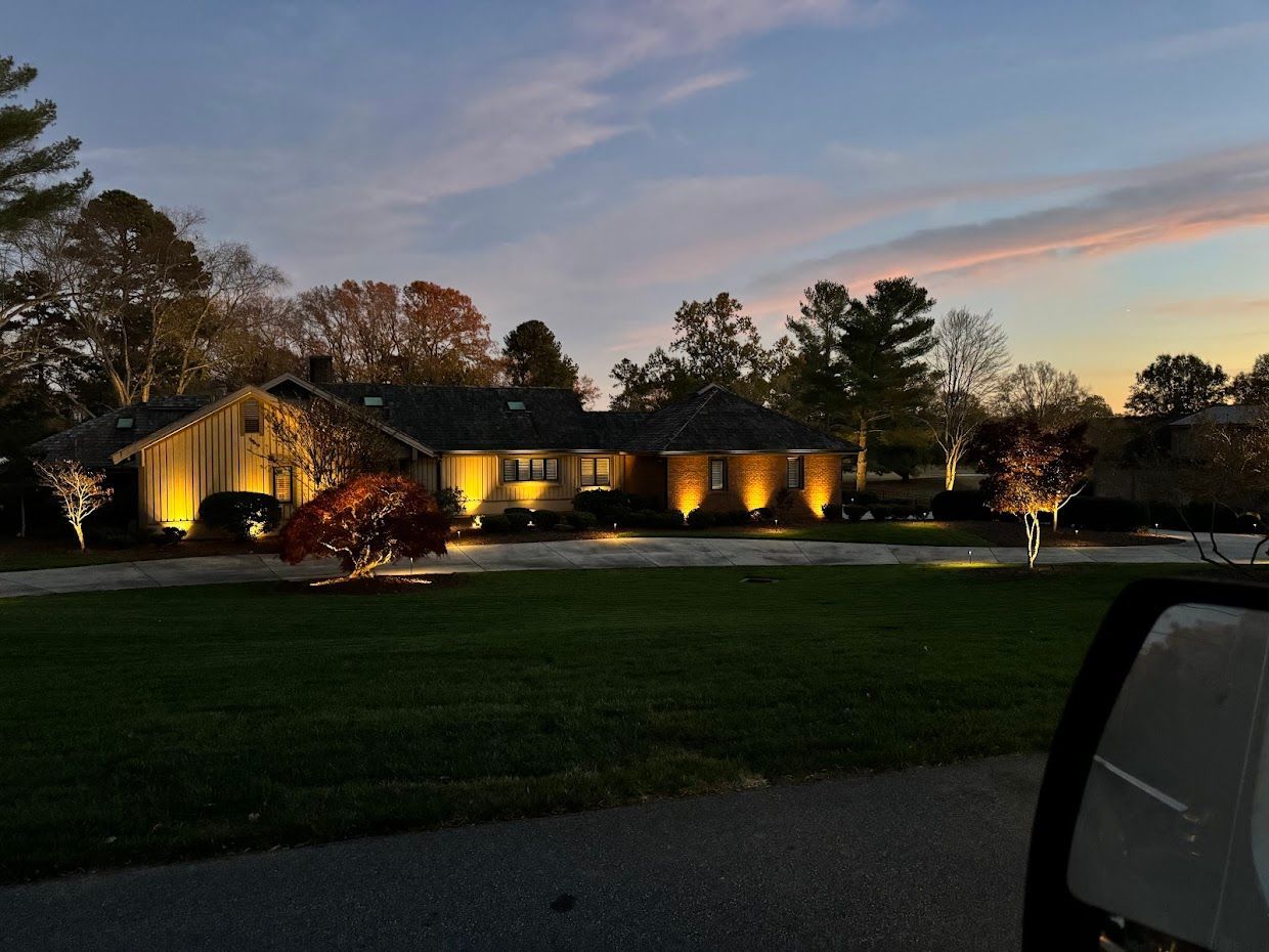 House at dusk lit with warm spotlights, trees and lawn in the foreground, cloudy sky.