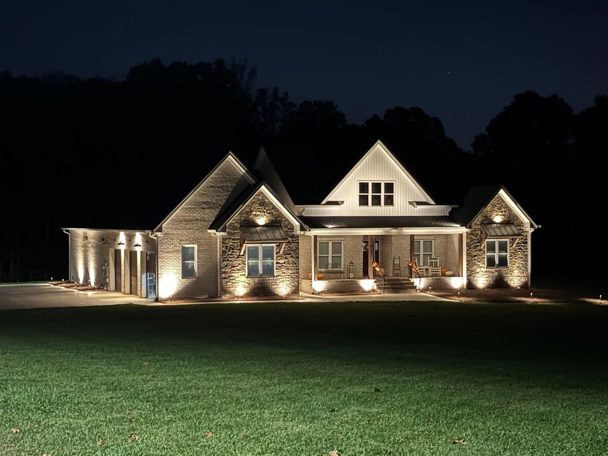 A house lit up at night, with landscaping lights illuminating the brick exterior and front porch, set against a dark background.