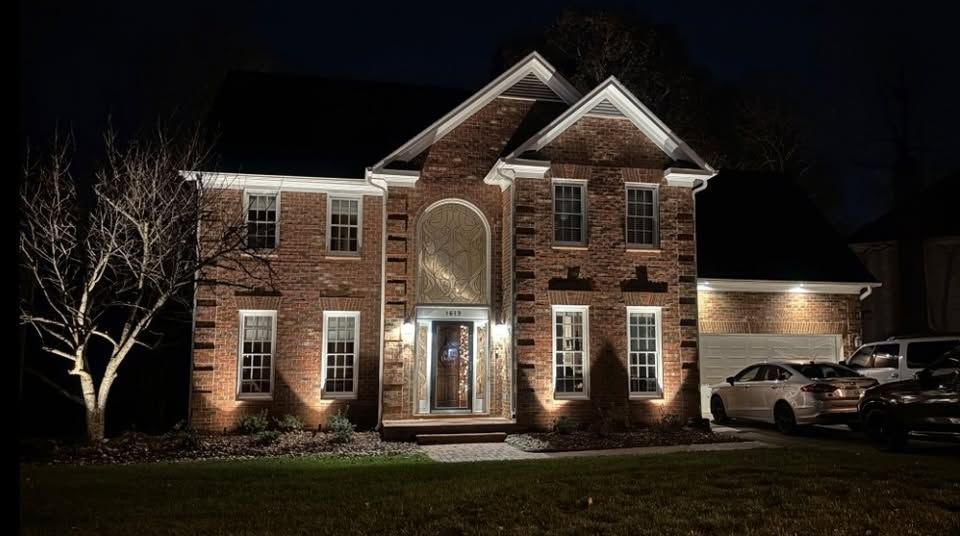 Brick house illuminated at night, with landscaping lights and cars in the driveway.