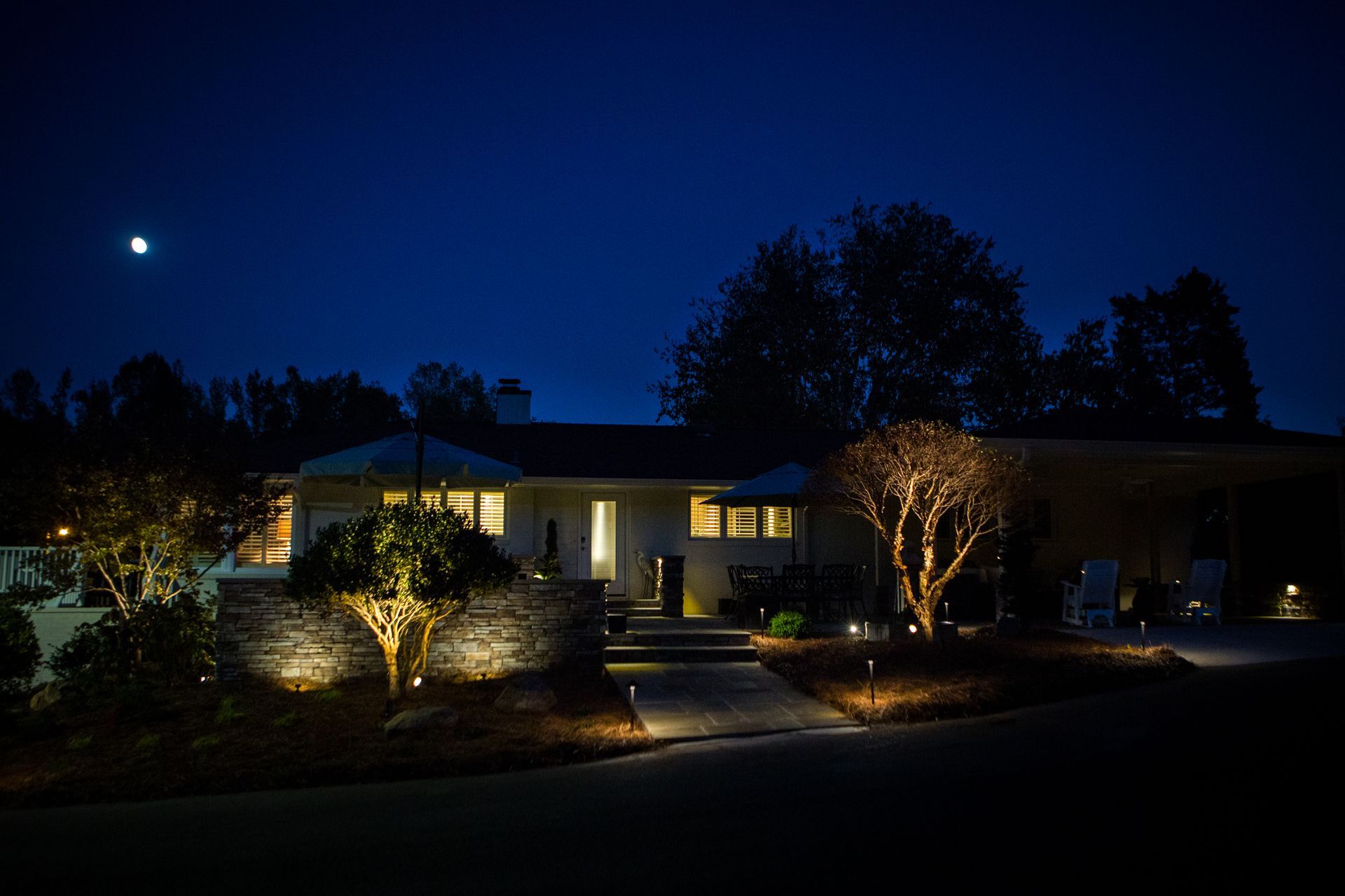 A house at night with landscape lighting illuminating the front yard, trees, and path.