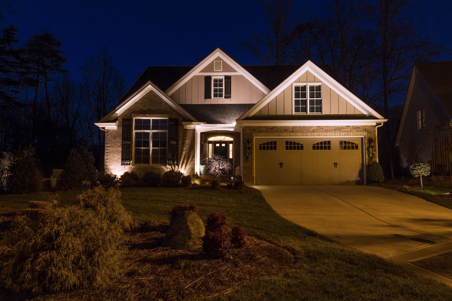 House exterior at night, lit by landscape lights. Brown siding and tan garage door on a dark blue background.