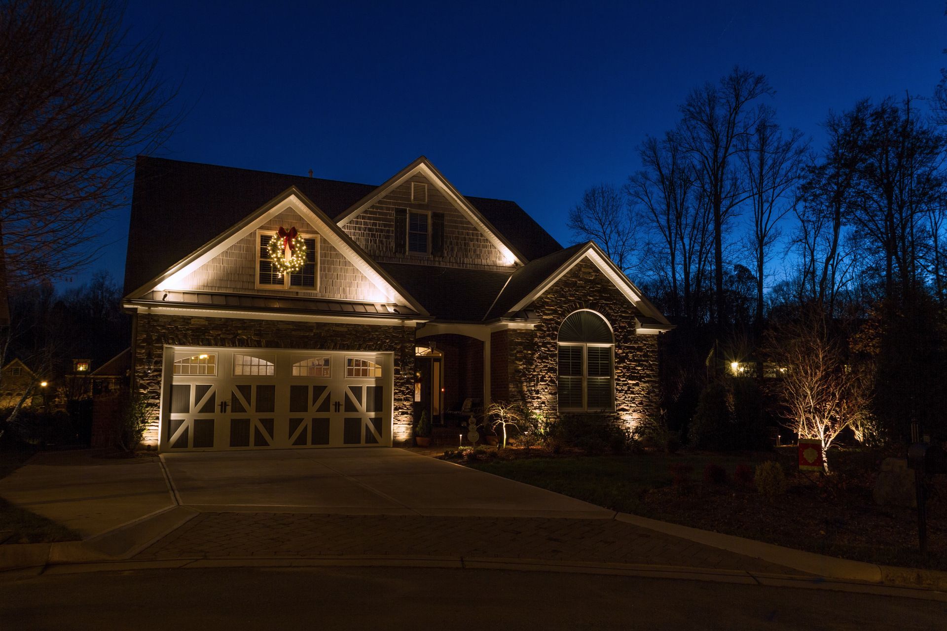 House illuminated at night with bright spotlights. Dark blue sky, Christmas wreath, and trees.
