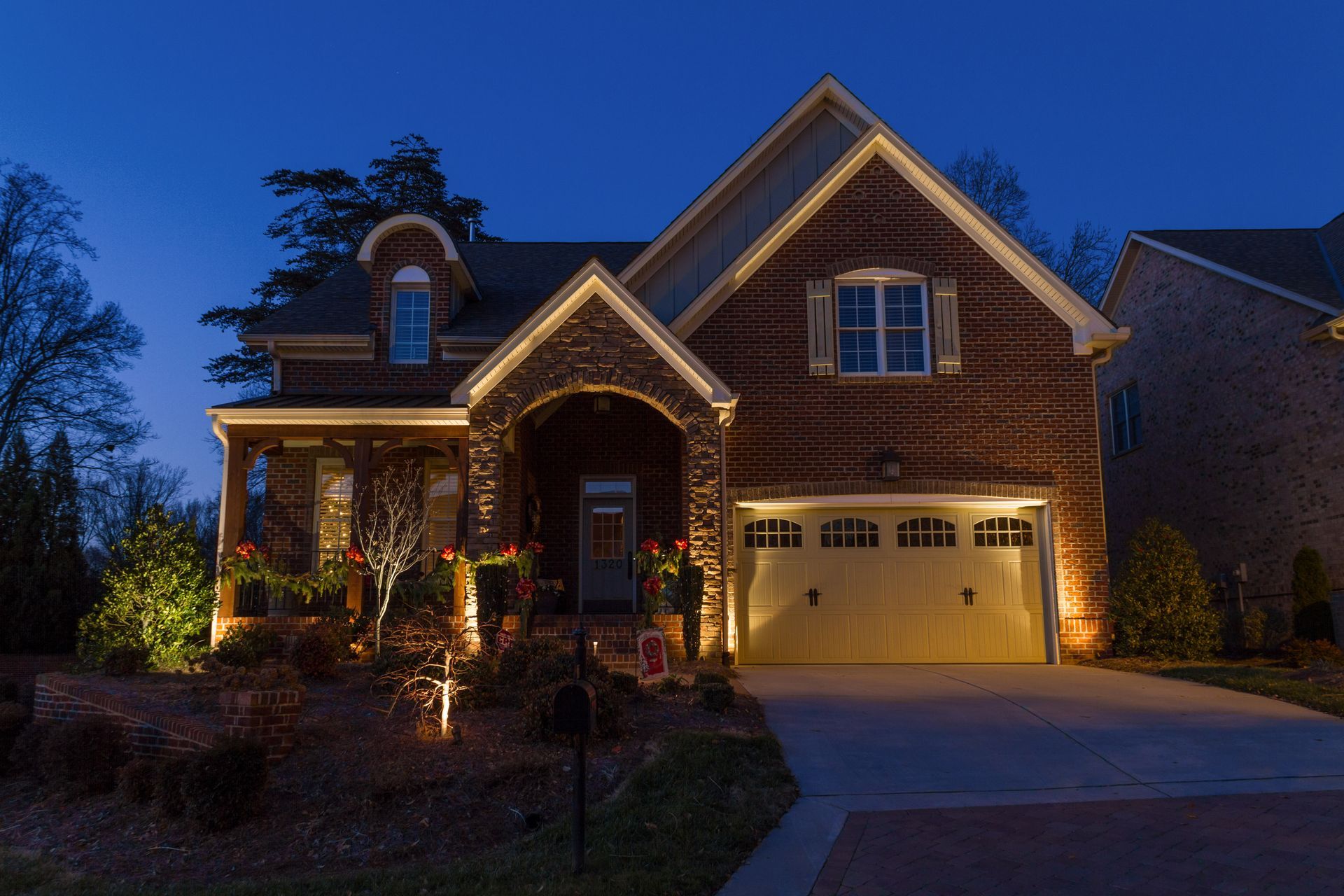 Two-story house illuminated at night with warm lights on the facade, porch, and landscaping; a dark blue sky.