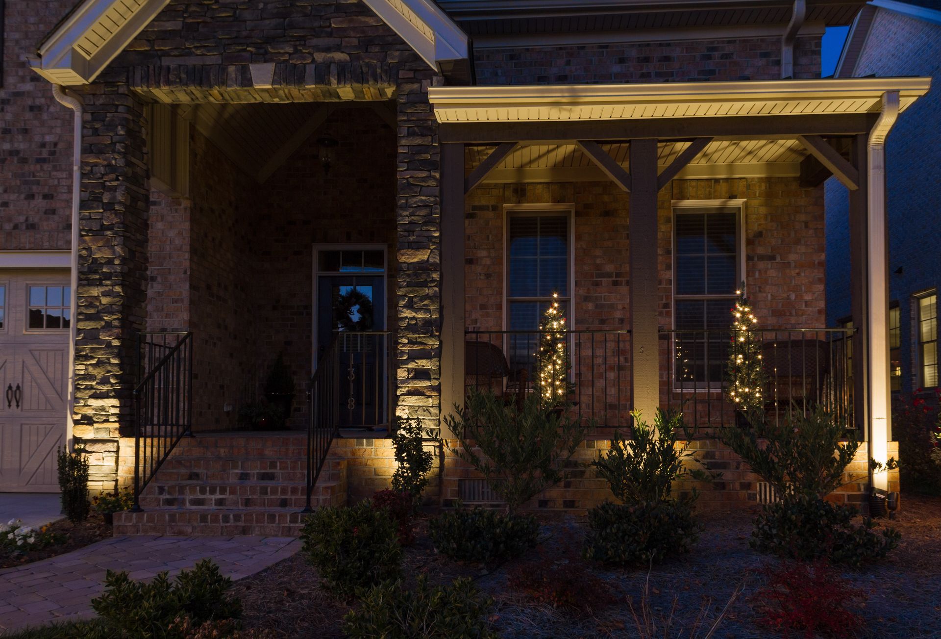 Night exterior of a house with lit porch and landscaping. Brick and stone facade, warm light illuminating the entryway.