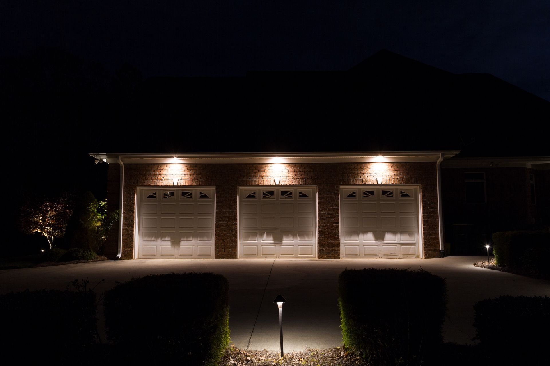 Lit garage with three doors, brick exterior, and landscape lighting at night.