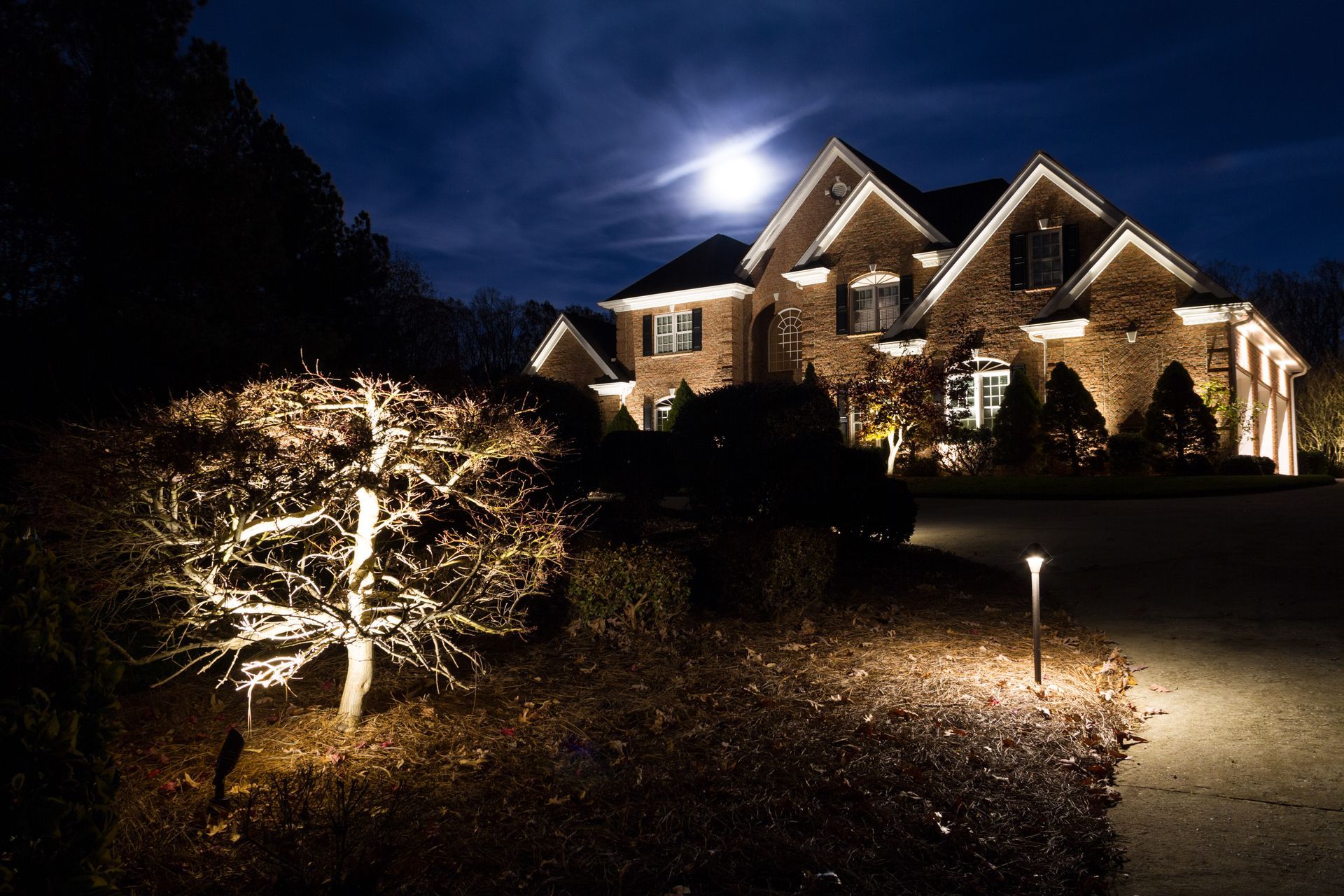 Lit-up house at night with spotlighted landscaping and a full moon in the background.