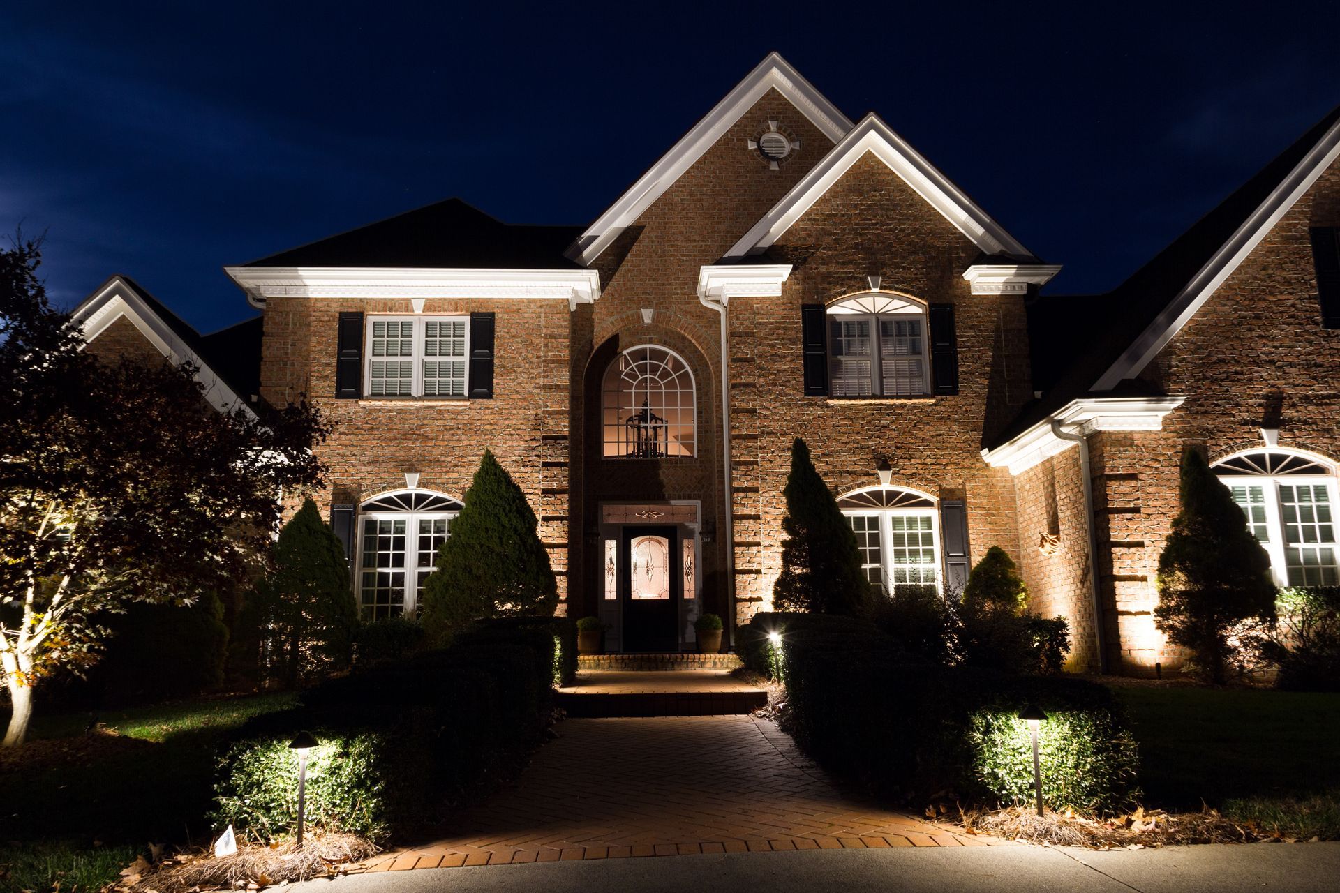 Brick house at night, illuminated by landscape lighting. Driveway, shrubs, and windows visible.