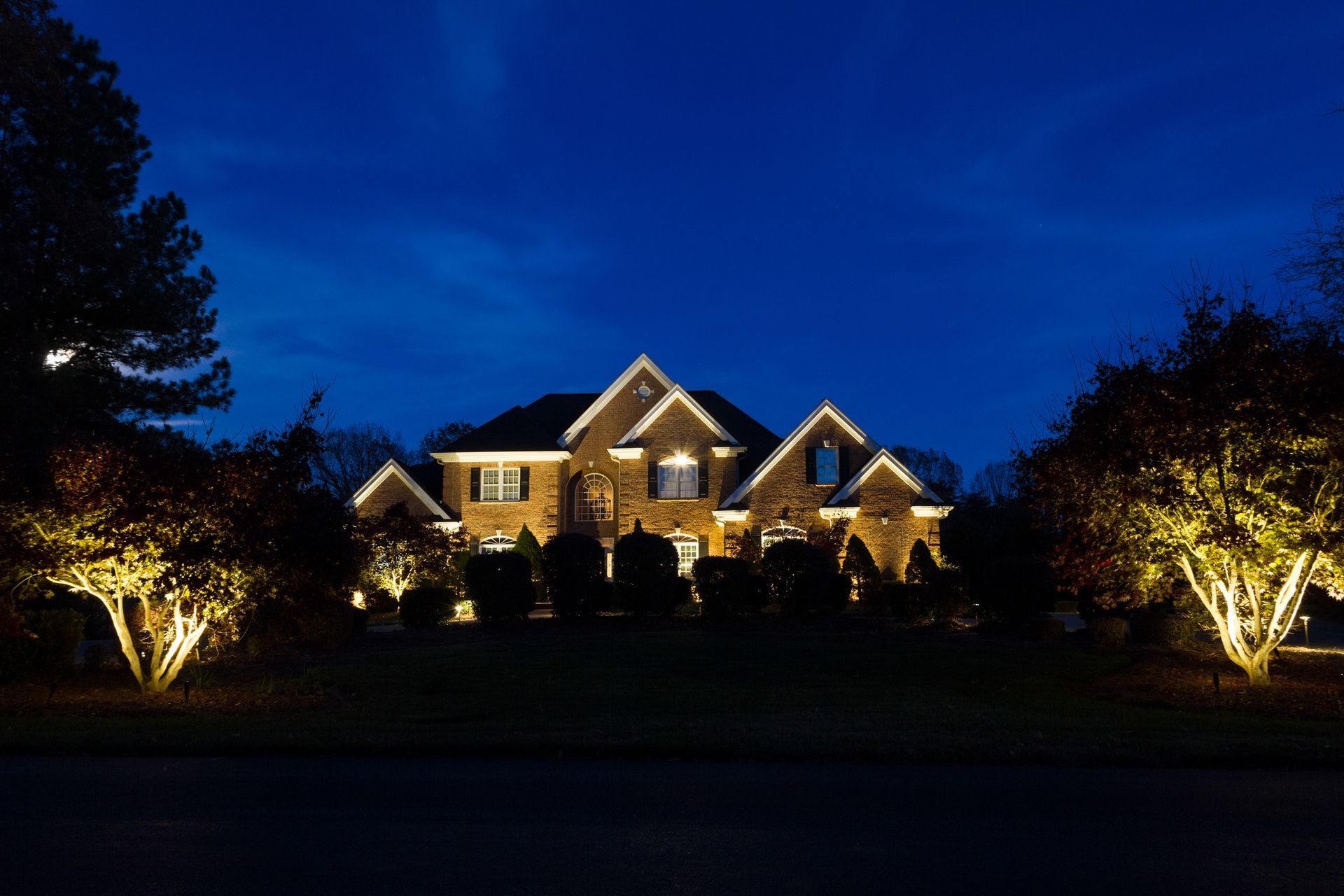 Lit house at night, illuminated by golden lights, framed by lit trees against a dark blue sky.