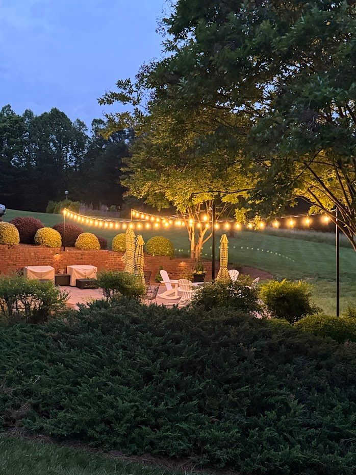 Patio with string lights at dusk; chairs, bushes, and a lawn.