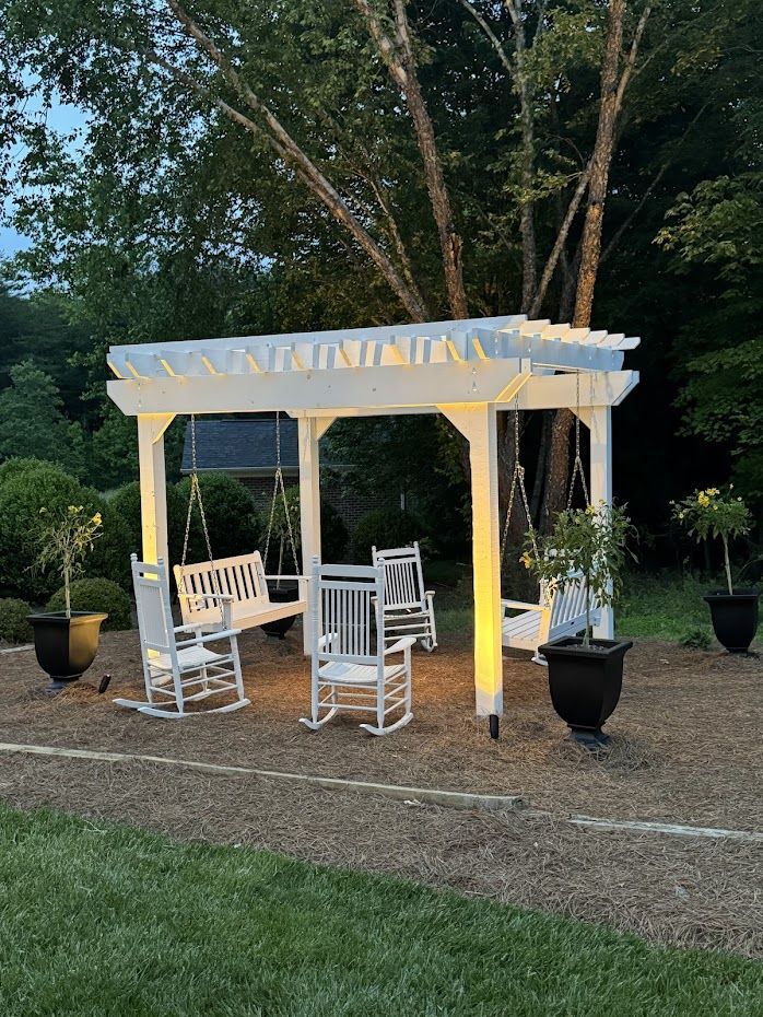 White pergola with swings and rocking chairs lit by overhead lights in a yard with potted plants.